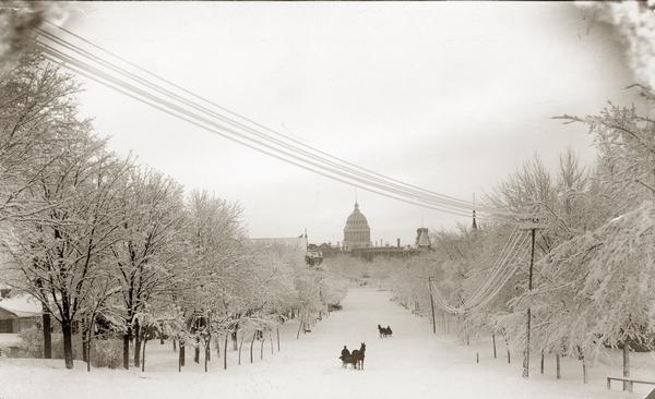 Winter scene looking down snow-covered Wisconsin Avenue to the third Wisconsin State Capitol. There are two horse-drawn sleighs, each with one passenger, on the street.