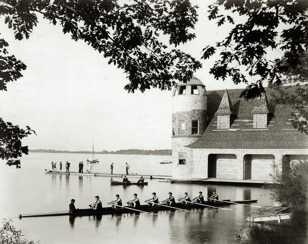 View from shoreline of a crew rowing a Pabst Shell on Lake Mendota near the University of Wisconsin-Madison boathouse, with two men in a canoe and people standing on the dock in the background.