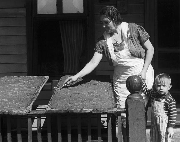 Loretta Falci (Lorenzina Fiocco Falci), an Italian woman, preparing sarsa, tomato paste on boards on the porch of her home, 622 Milton Street, as she holds the hand of a young boy (Joseph Anthony Pellittei). Milton Street is located in what was known as the Greenbush neighborhood.