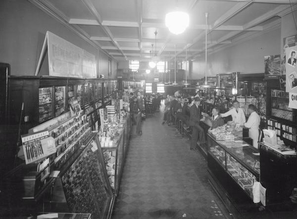 Customers pose at the soda fountain counter. Clerks are behind the counter.