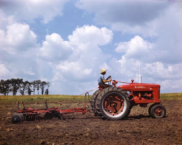 Color photograph of a farmer with a straw hat working a field using a Farmall M tractor with disc harrow against a bright blue sky strewn with fluffy white clouds. The photograph was most likely taken at or near International Harvester's experimental farm in Hinsdale, Illinois.