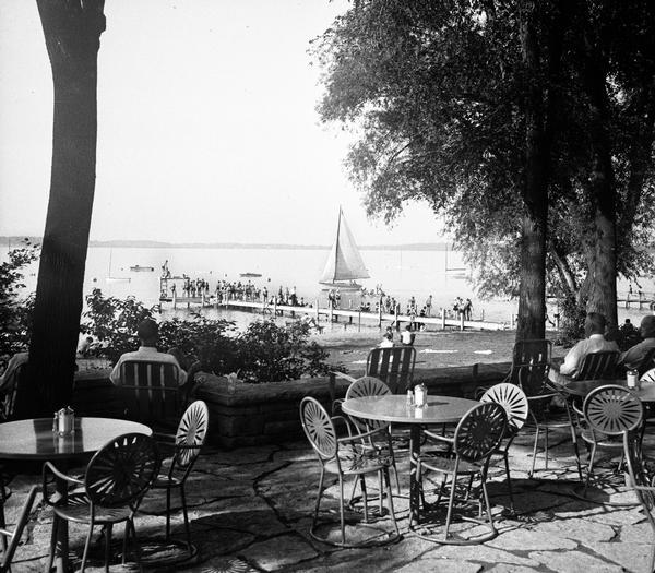 University of Wisconsin-Madison Memorial Union Terrace, with tables and chairs, looking out over Lake Mendota. There is a pier and a sailboat on the water.