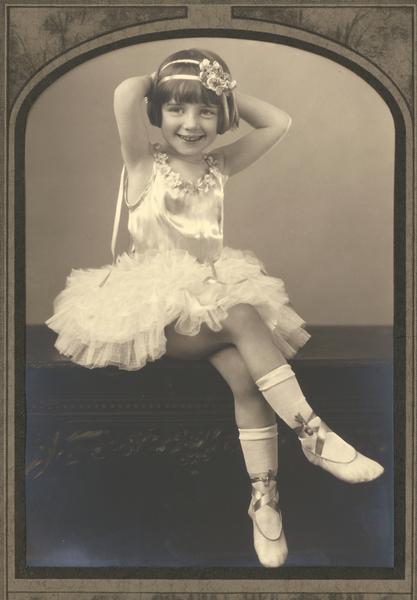 Young ballerina of the Kehl School of Dance poses in her dance costume which was hand-designed by Kehl seamstresses. Satin ribbons tied around the ankles and socks, instead of tights, were the norm at this time.