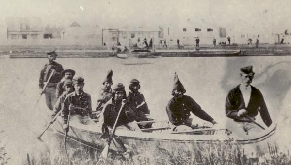 Nine men arrive at Fort William by canoe. Fort William, also called Kaministiguia, was the Hudson's Bay fur trading post. Men are standing along the opposite shoreline in front of buildings.