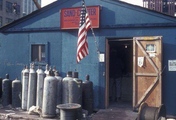 Office for the Sand Chapter of Mechanical Contractors at the construction site for the World Trade Center. The construction office has an American flag hanging by the front door and there are several acetelyne tanks to the left of the door.