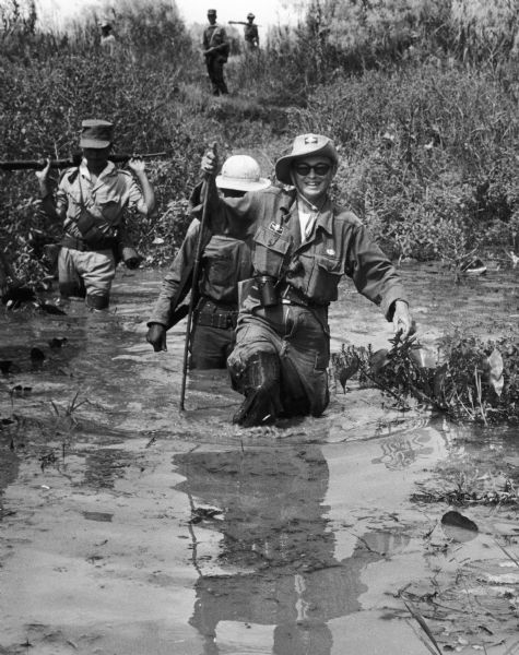 Dickey Chapelle dressed in military clothing wading through a swamp in Vietnam.