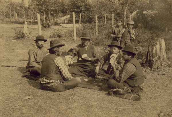 Five Winnebago men are sitting on the ground around a blanket playing cards. Two young boys are standing behind them watching.