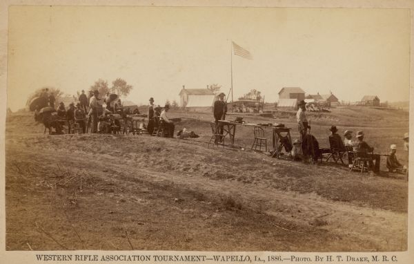 Men and boys participate in the Western Rifle Association Tournament.  Many of the participants are sitting under umbrellas and holding their guns.  Several tents of spectators are in the background.