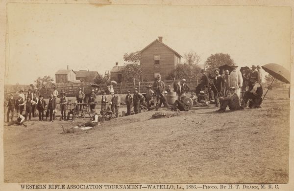 Participants at the Western Rifle Association Tournament in Wapello, Iowa.  Many of the men are holding guns.  A farmhouse and farm buildings are in the background.  Several of the participants are sitting under umbrellas.
