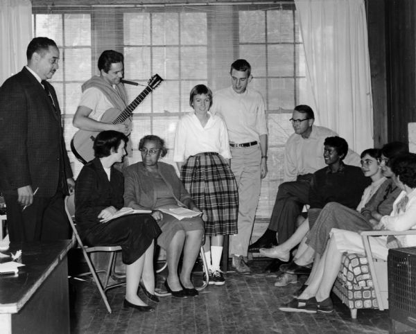 Eleven people, including Thurgood Marshall, Anne Braden, Myles Horton, and Septima Clark, are sitting and standing during a Civil Rights meeting at Highlander School. Guy Carawan is holding a guitar.