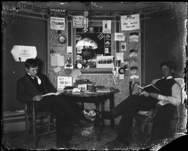 Two men read in a room with many signs on the wall and a hanging lamp. Perhaps a fraternity house.