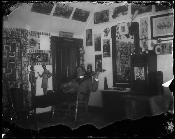 Man in dormitory or fraternity playing banjo. There are signs that say "University of Missouri" and "Don't be woozy" above the door. There are photos and cards posted on the wall.