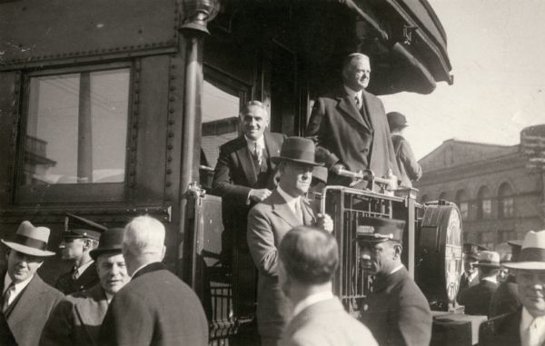 Herbert Hoover stands with Governor Kohler on the back of a train during a campaign visit. The man in the hat holding the rail of the train car is U.S. Secret Service agent Richard Jervis.