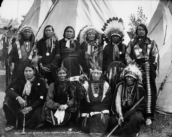 Outdoor group portrait of men, with tipis behind them. Caption reads: "Group of Sioux and Apache Indians." Identifies Geronimo, first row, second from right, a chief of the Chiricahua Apaches.