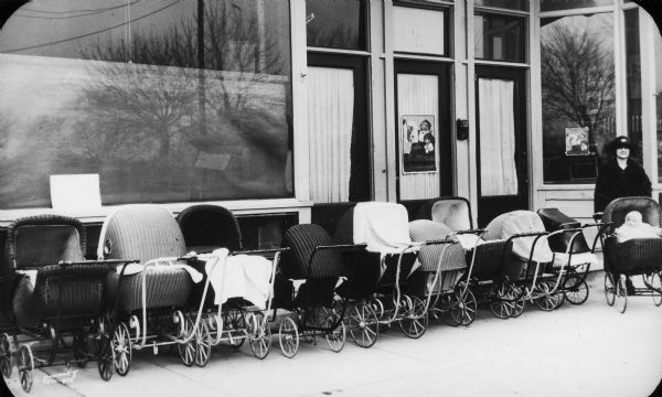A mother and infant in a baby carriage pose at the end of a row of wicker baby carriages in front of what is probably the Neighborhood House.