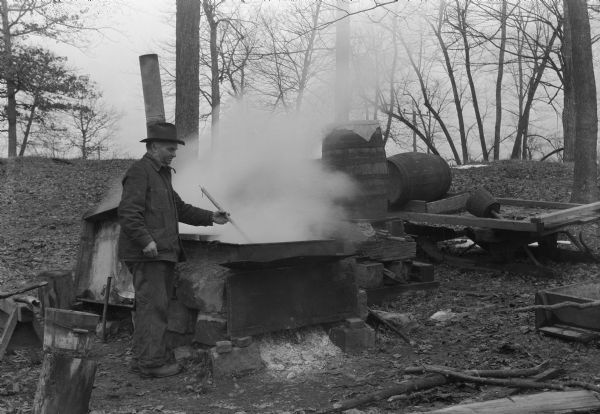Man in "sugar bush" (grove of maples) boiling down maple sap in production of maple syrup. Probably Wisconsin.