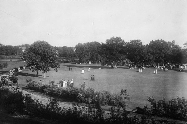 Elevated view of Vilas Park, filled with groups of people on the lawn, and horse-drawn carriages on the drive.