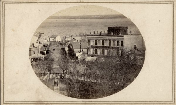 Intersection of King and East Main streets photographed by John S. Fuller from the Wisconsin State Capitol roof, about 1860-1863.  In the foreground two strollers can be seen leaving the Capitol Park while Lake Monona is in the distance.  The large building on the right is the Fairchild Block which was constructed by Jairus Fairchild in 1853.