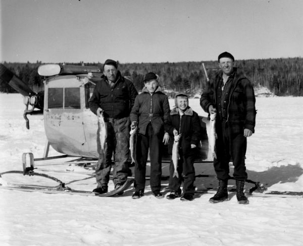 Two men and two boys standing on ice in front of a windsled. All four are holding lake trout.