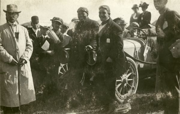 African American heavyweight boxing champion, Jack Johnson, drove up to Milwaukee from his home in Chicago to watch the Vanderbilt Cup race on October 2, 1912. A number of unidentified people are gathered around him as he and another man (both wearing goggles) lean against a car (probably Johnson's).