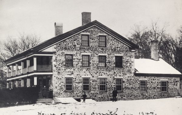 Side view of the Jesse Smith tavern, built in 1847. Two people are standing in front of the tavern in the snow. Handwriting at the bottom reads: "Home of Jesse Smith in 1847".