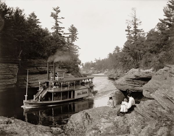A woman holding a baby sits near a little girl on rocks looking down on the "Dell Queen" steamboat entering the Narrows at Devil's Elbow.