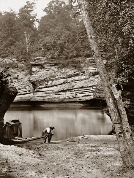 View up the river from behind Gates Ravine. There is a boat and dark tent at the edge of the river. H.H. Bennett is sitting on rowboat with his wetplate tent behind him.