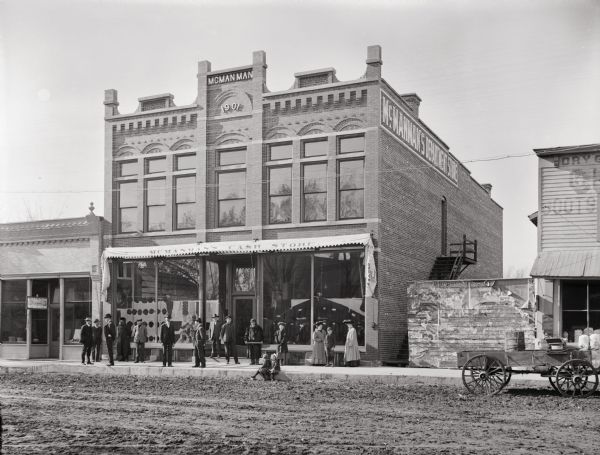 External view of McManman Store, Superior Street, Kilbourn, with people posing standing in front of the store, and two children sitting on the curb.