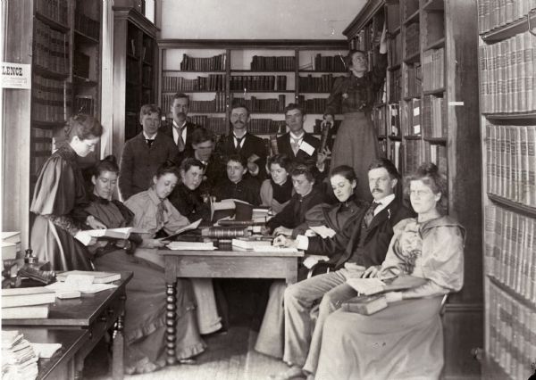 Frederick Jackson Turner with his History Seminar in an alcove of the Historical Society rooms in the third Wisconsin State Capitol, about 1893-1894. Back row, from left: W.B. Overson, J.M. Johnston, Joseph Schafer, Charles L. Baldwin, and Florence Baker Hayes (staff).  Front row, from left: Emma Hawley (staff), Estelle Hayden, Kate Bucknam, Ada Taylor, Annie Pellow, Dena Lindley, Sadie Bold, Flora Barnes, Turner, and Catharine Cleveland. The young man in the center, bending over, is E.F. Dithmar.