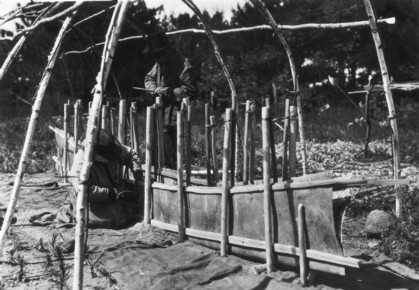 An Ojibwa man shaping birch bark in a frame during the process of making a canoe.