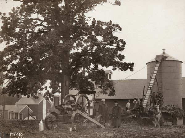 Early International Harvester engine, probably a Titan, powering an elevator to lift silage into a silo.  Two men in overcoats and two children in overalls look on.