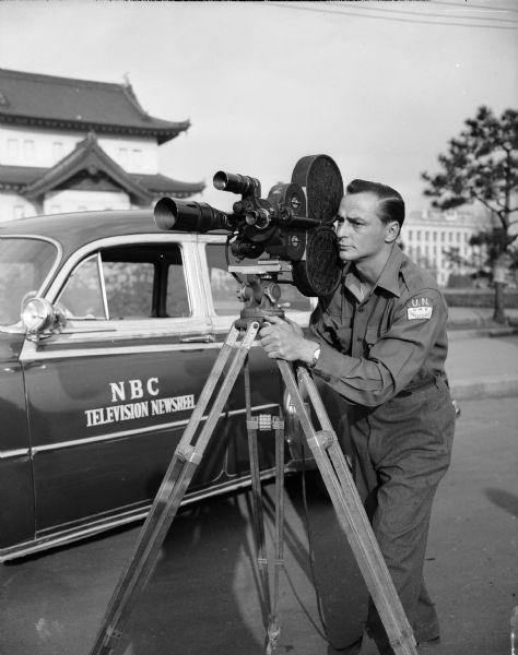 A U.N. war correspondent is shown filming with a news camera next to an NBC Television Newsreel vehicle.