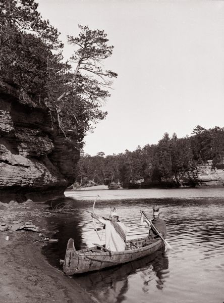 Two men dressed as "Indians" are sitting in a canoe at the edge of the beach. One is posing holding a bow and arrow, the other is holding a paddle.