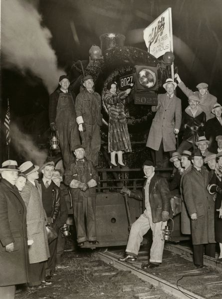 Schlitz train and group of revelers, celebrating the end of prohibition in Milwaukee. Schlitz Engine #8027 departs to deliver the first Schlitz beer at 12:01 a.m. on April 7, 1933. Pictured from left are R.D. Miller, Superintendent; (on front footboard) Jesse Worner, Yardman; (in front of draw casting) A. Basta, Fireman; Edward Wroblewski, (on running board above front beam) Anton Plewa, Conductor; (between Conductor and woman) and William H. Kay, Engineer.