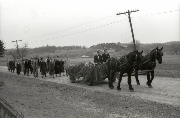 Funeral procession of Frank Lloyd Wright to Unity Chapel Cemetery. His body is carried in a horse-drawn vehicle with a group of people marching behind.