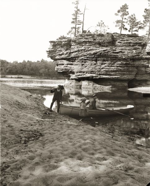 View down sandy beach towards H.H. Bennett and Ruth Bennett on shore near Sugar Bowl. H.H. Bennett is holding a camera over his shoulder. There is an unidentified man in a rowboat at the shoreline.