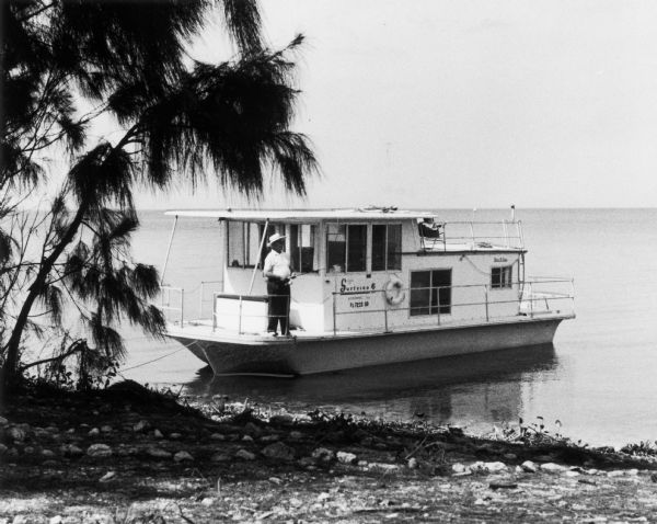View from shoreline towards a man standing on the deck of a houseboat, which has the name "Surfside 6" on its side.