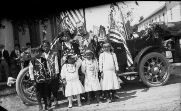 An Ottawa Indian family in traditional dress pose next to a car with a Caucasian man. The car is decorated with American flags as though for a special occasion.