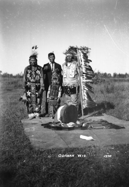Native American family, dressed in native clothing, standing on blanket outdoors.