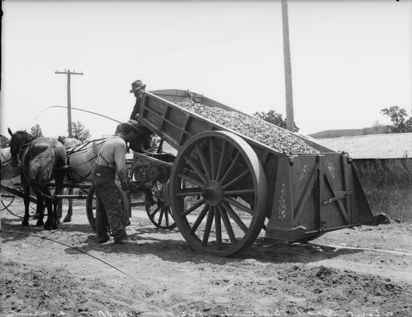 Two men spreading gravel from a horse-drawn dump wagon onto a road. The wagon was manufactured by Road Machine Co.