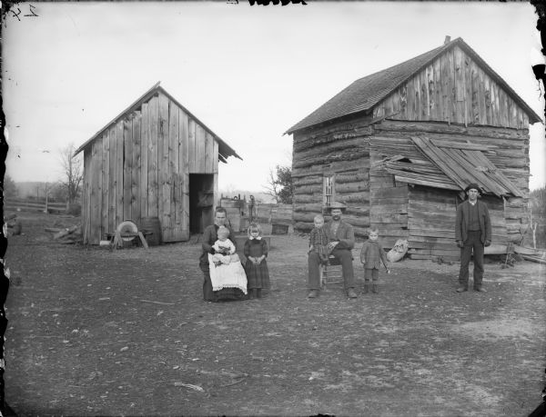 Man and woman posing sitting, each holding a child in their lap, and a young man, girl, and a boy holding a toy horn, are posing standing. The group is posing in the yard in front of a log house and farm building. According to Bonnie Evans of Cataract, Wisconsin, this is the Chas. Koehler family, and the man on the right is Gust Jones, a nephew.