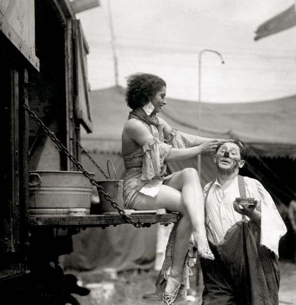 Circus performer Harriet Hodgini, sits on the gate of a truck helping Otto Griebling, a circus clown, apply his makeup.