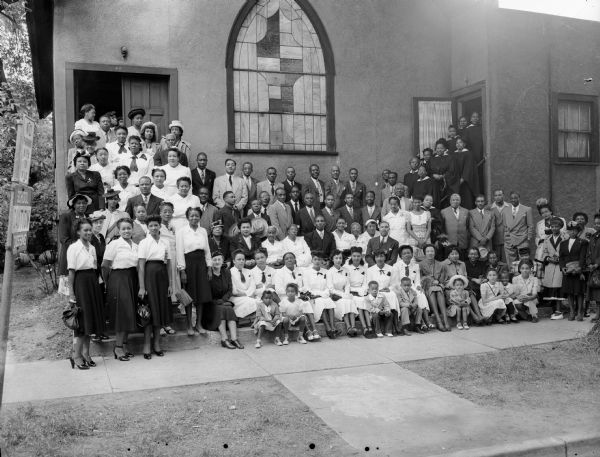 Group portrait of the Mt. Zion Baptist Church congregation in front of the church, 548 West Johnson Street, with pastor Rev. Cunningham.  Members are primarily African-American.
Children in the front row: Beatrice and Jeanette Braxton, Richard Cunningham, and two of the Taliaferro daughters.  The first row of women seated include: Mrs. Ross, Mrs Cunningham, Dorothy McKinley, Charlie Allen, Addrena Squires, Oletha Montgomery, Willa Jo Withers, Blanche Blue and Anna Banks. The choir to the left include: Gertrude Taliaferro, Ola Jordan, Bertean Green, Essie Dunn, Mabel Marel, Jewel Mosley, Mary Hargrove, Emmette Williams, Maggie Davis, Mary Lee Martin, Gertrude Dixon, Dale Craven, Mary Calvin, Essie Jones, Eunice Guy, Arlene Taylor, and Wilma Mae Taylor.
Members in second row include: Olga Jordan, Lujuna Grandison, Mrs. Joseph Washington, the Rev. Cunningham, Mrs. Matthews, Mother Hodge, Marg Jones, the Rev. Joseph Washington, Mrs. Battiste, Rosie Lee Matthews, Mr. Orange, Alex Benford, Arthur Walker and J.C. Calvin.
Third row: Bill Martin, Larry Adams, Richard Sims and Mr. Guy.
Back row includes Jesse Guy and Roosevelt McKinley.
The choir in the background is believed to be guests from out of town.

