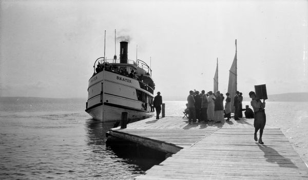 A group of people gather on Mission dock as the steamboat "Skater" approaches. A boy is walking down the dock carrying a box on his shoulder.