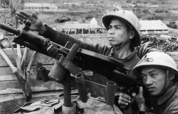 Two North Vietnamese soldiers pose with machine guns ready to fire. The caption indicates that the men are workers at an electric factory using machine guns to bring down U.S. planes in North Vietnamese airspace.