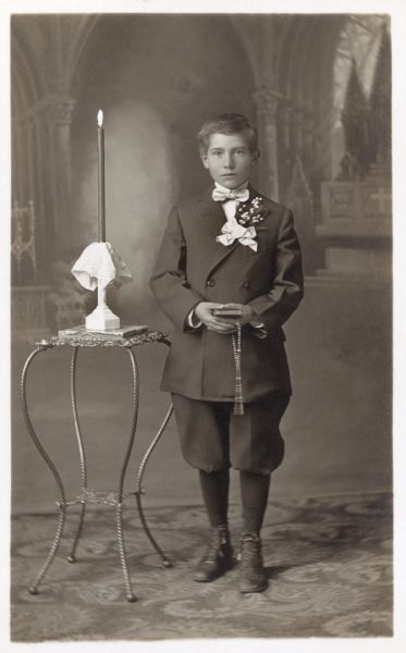 Photographic postcard of a studio portrait of Carl Virnig on the occasion of his First Communion. He is dressed in a jacket and knickerbockers (knickers) and holds a rosary and catechism. He is posed in front of a painted backdrop.