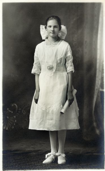 Photographic postcard of a full-length studio portrait of Lanona Petersilie Wipperfurth on the occasion of her graduation. She is wearing a white dress, a necklace, and white stockings and white shoes with bows. She has her hair pulled back and a large bow in her hair, and is holding a rolled diploma in her left hand. She is posing in front of a painted backdrop.