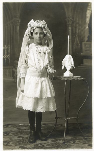 Photographic postcard of a full-length studio portrait of a girl on the occasion of her First Communion. She is standing and wearing a white dress, flowered tiara with veil, a crucifix on a necklace, and dark stockings and shoes. She is holding a catechism and rosary in her left hand. She is posing next to a table with a candle in a candle holder in front of a painted backdrop.