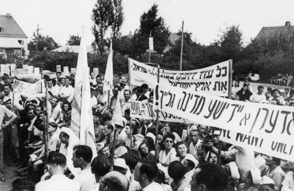 Refugees demonstrate for Israeli statehood at a Displaced Persons camp; Germany.

Saul Sorrin was interviewed as part of the Wisconsin Survivors of the Holocaust Interviews project. Sorrin, born in New York in 1919, applied in 1940 for a position with the United Nations Relief and Rehabilitation Administration (UNRRA). He worked with Holocaust survivors as a supply officer for UNRAA team 560 at the Displaced Persons camp Neu Freimann Siedlung in Germany and later, at General Dwight D. Eisenhower's recommendation, Sorrin became the Area Director of the International Refugee Organization based at the Wolfratshausen DP camp in Bad Kissingen.

When asked about this image, Sorrin revealed, "At the time that the United Nations was considering the partition, when the debates were going on, or at times when the United States was getting ready to make some statement, or whatever, at certain political moments, the Jews of the camps were politically quite active and would engage in demonstrations for Israel. That's what it really was all about. And this is one of these demonstrations. I can see here in Yiddish, "Mir fodern a Yidishe medine [blaykh?], "We demand a Jewish state now." And signs and placards were made. It's possible that there was a visitor, a commission of some sort may have visited one of the camps.  The Anglo-American Commission of Inquiry or United Nations Commission of Inquiry, whatever. But demonstrations and political activity were very, very much a part of--when I say political, I mean in a Zionist sense--were a part of life in the Jewish camps."

Interview by Jean Loeb Lettofsky and David Mandel, March 3, 1980.