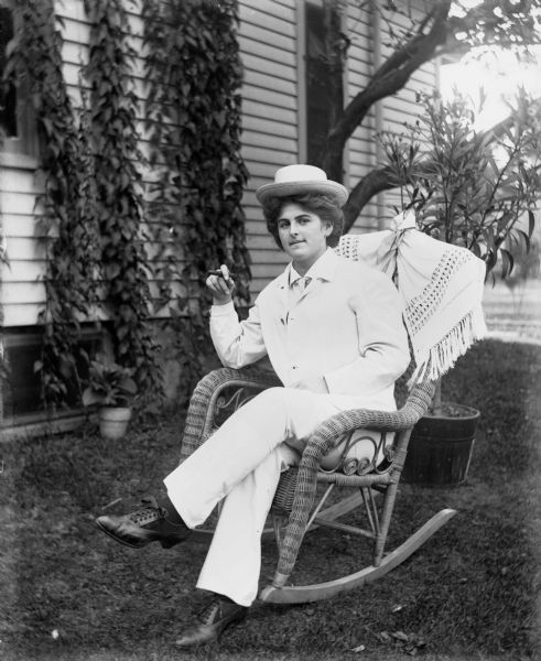 Outdoor portrait of Hattie Fels Owen wearing a white suit and hat and sitting in a wicker rocking chair. She is holding a cigar in her right hand. Behind her ivy is growing along the siding of a house. Potted plants are on the lawn.